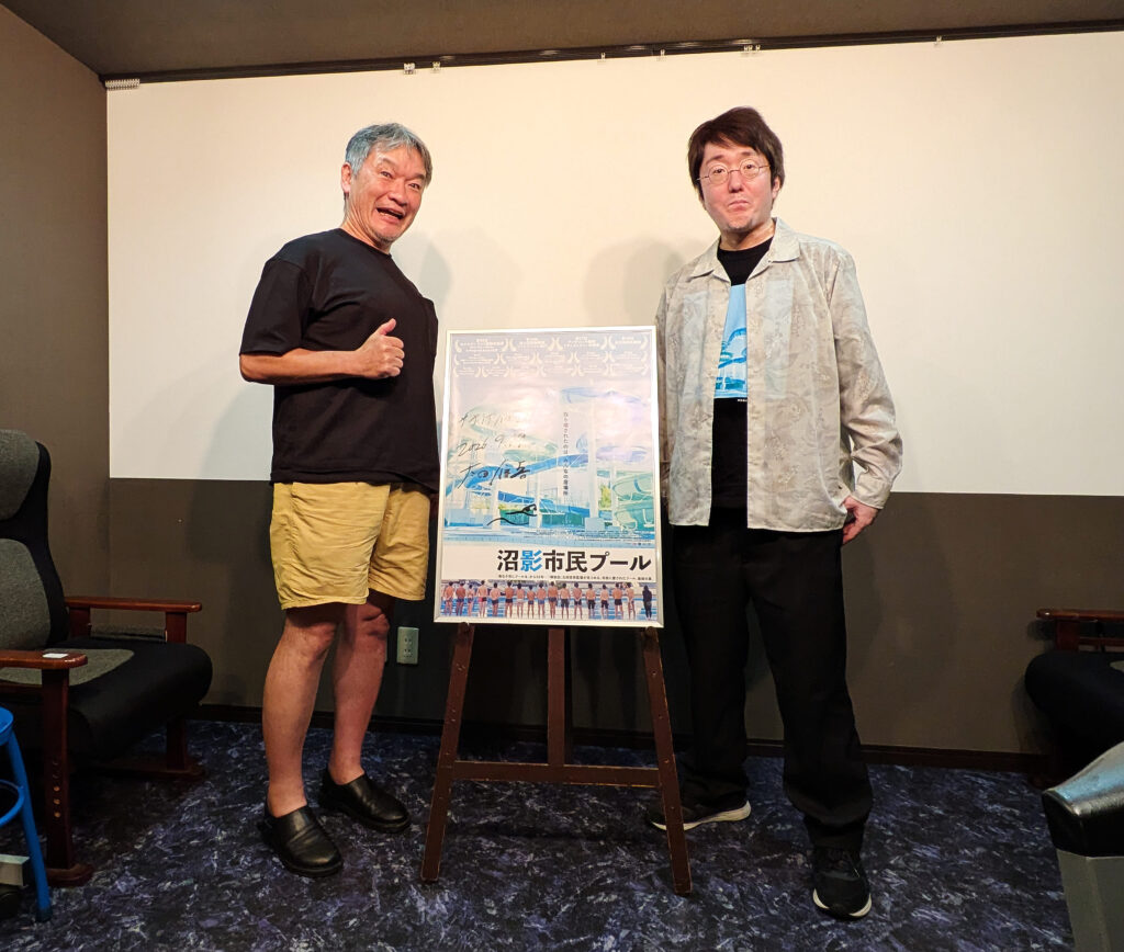 映画『沼影市民プール』のポスターを挟んで立つ太田信吾監督とインタビュアー