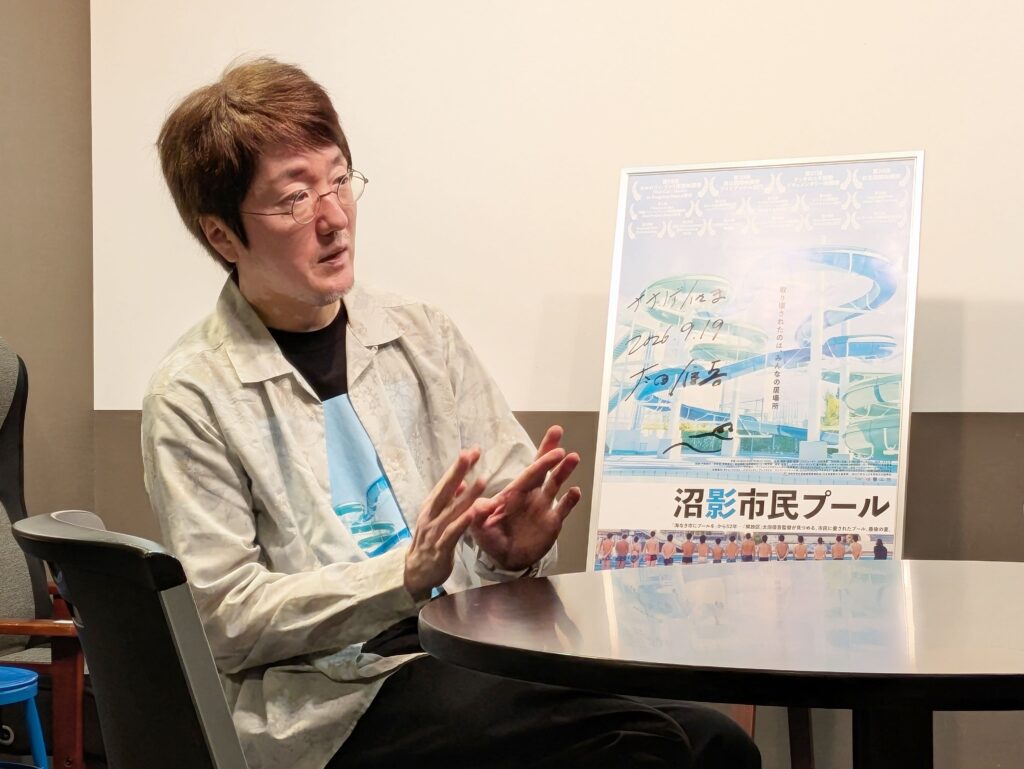 映画『沼影市民プール』のポスターを前にインタビューに答える太田信吾監督
